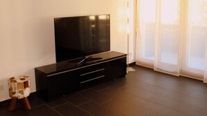 Ein modernes Wohnzimmer mit einem Flachbildfernseher auf einer schwarzen Unterhaltungsanlage, einem kleinen Holzschemel und einer Glastür, die auf einen Balkon führt.