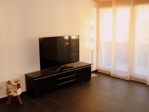 Ein modernes Wohnzimmer mit einem Flachbildfernseher auf einer schwarzen Unterhaltungsanlage, einem kleinen Holzschemel und einer Glastür, die auf einen Balkon führt.