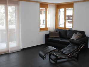 A modern living room with a black leather sofa, a black reclining chair, and large windows with wooden frames. - Weisshornblick, Sandhubel Appartement/Fewo, Bad, WC, Bergseite Appartement/Fewo by Interhome (Arosa)