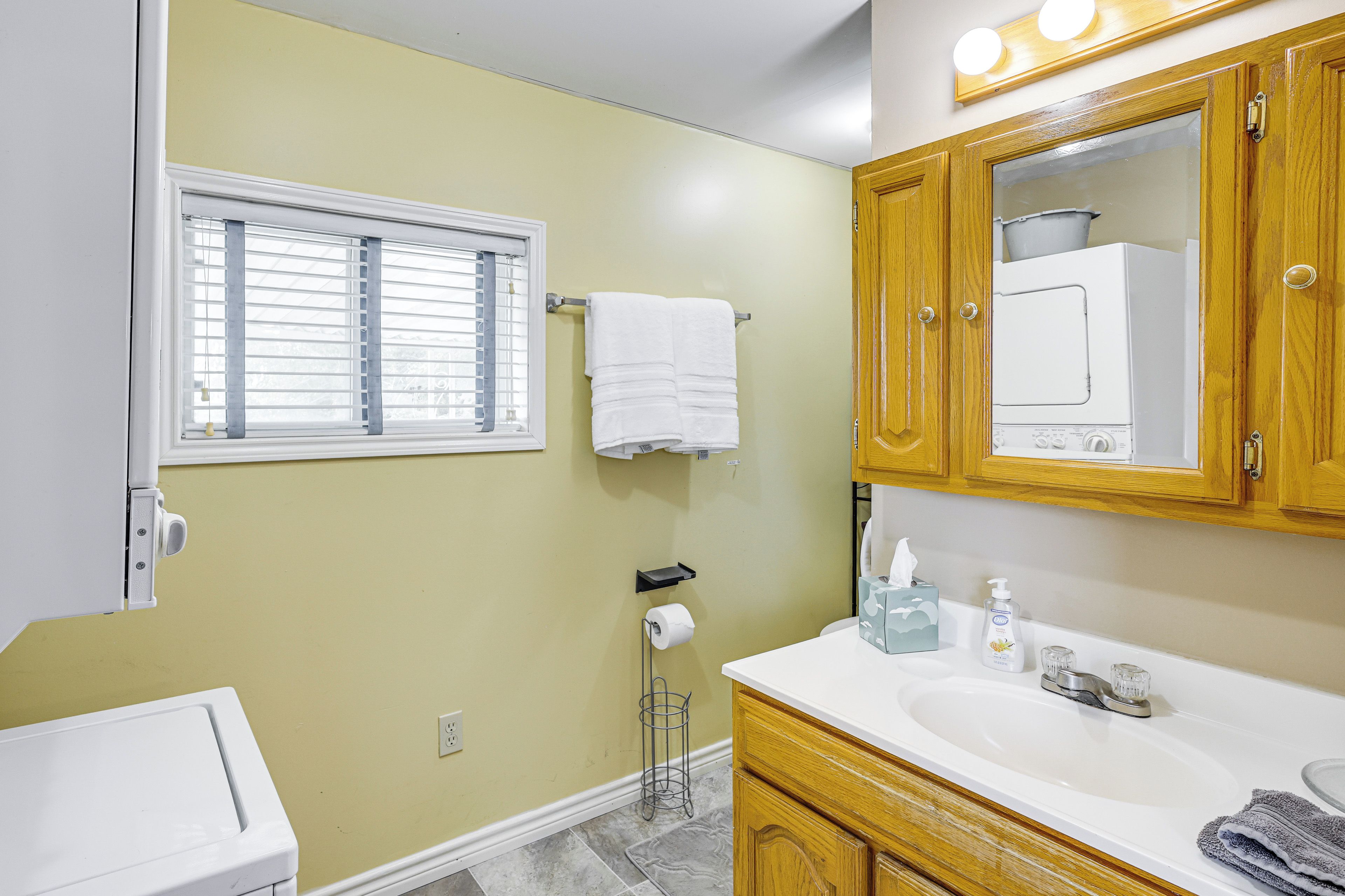 A bathroom with a white sink, wooden cabinets, a mirror, and a window with blinds.