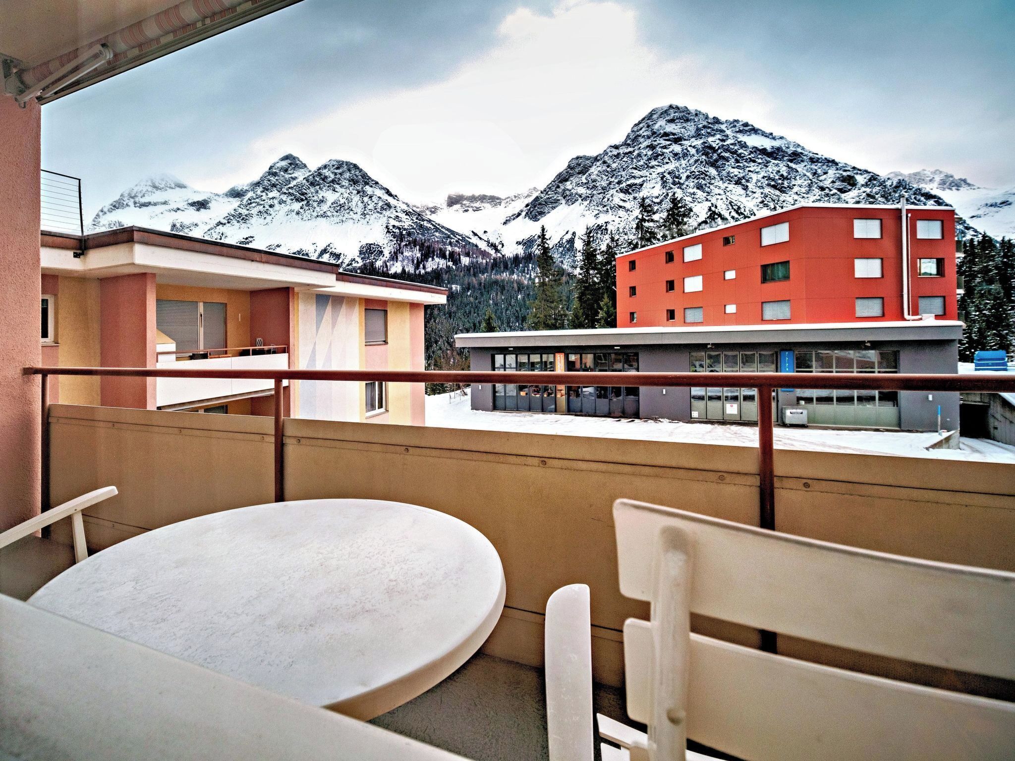 A balcony with a table and chairs, overlooking a building with a red facade and snow-covered mountains in the background.