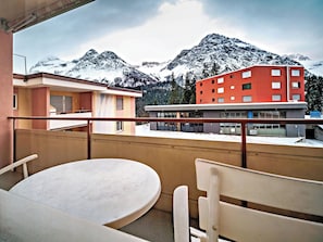 Ein Balkon mit Tisch und Stühlen, Blick auf ein Gebäude mit roter Fassade und verschneite Berge im Hintergrund.