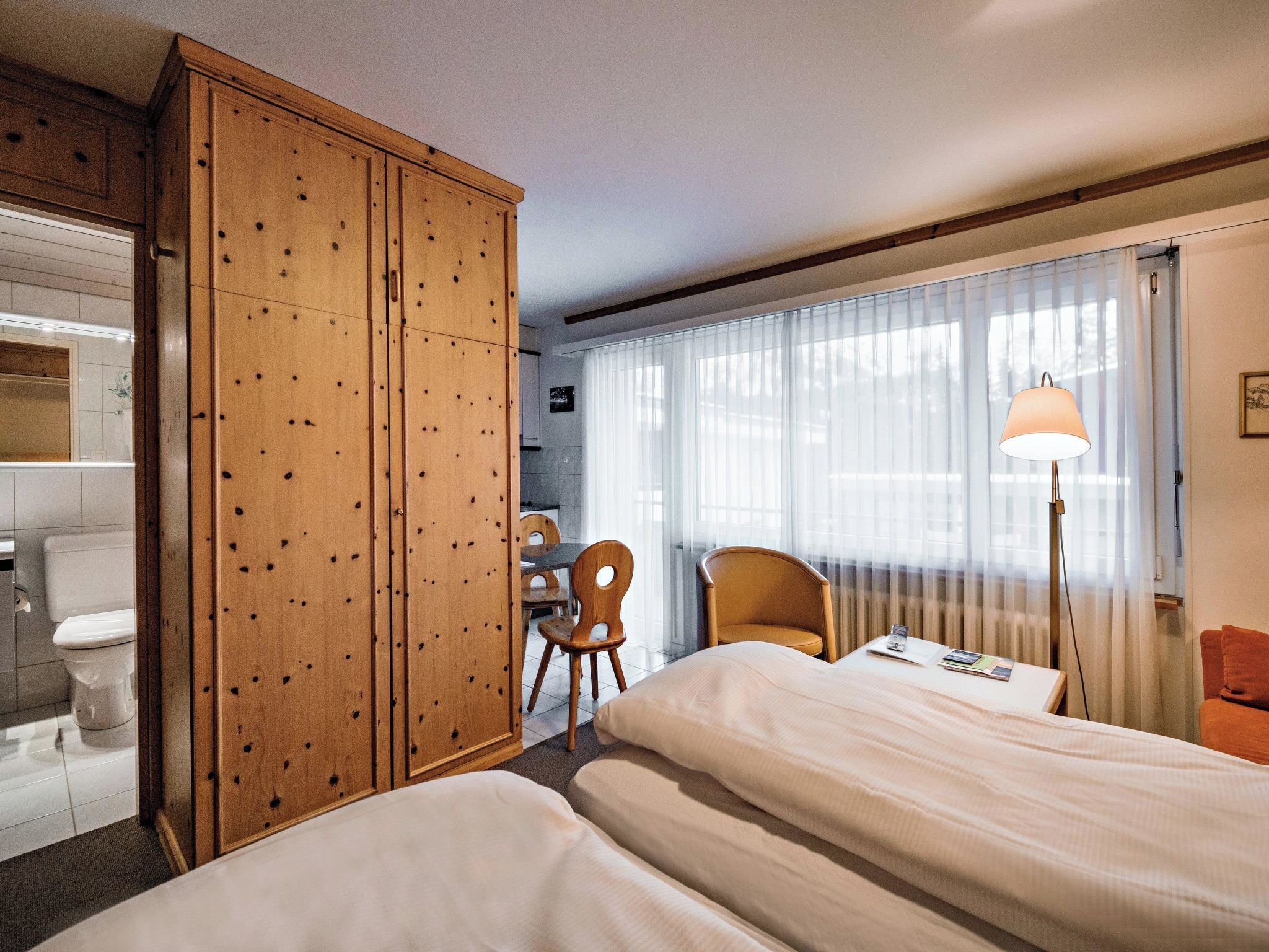 A hotel room with two beds, a wooden wardrobe, a desk with a chair, and a window with curtains.