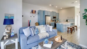 A modern living room with a sofa, coffee table, and kitchen area with blue cabinets and a central island. - Walk to Beach! North Padre Home w/ Pool Access (Corpus Christi)