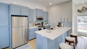 A modern kitchen with blue cabinets, stainless steel appliances, a central island with stools, and a view through a large window. - Walk to Beach! North Padre Home w/ Pool Access (Corpus Christi)