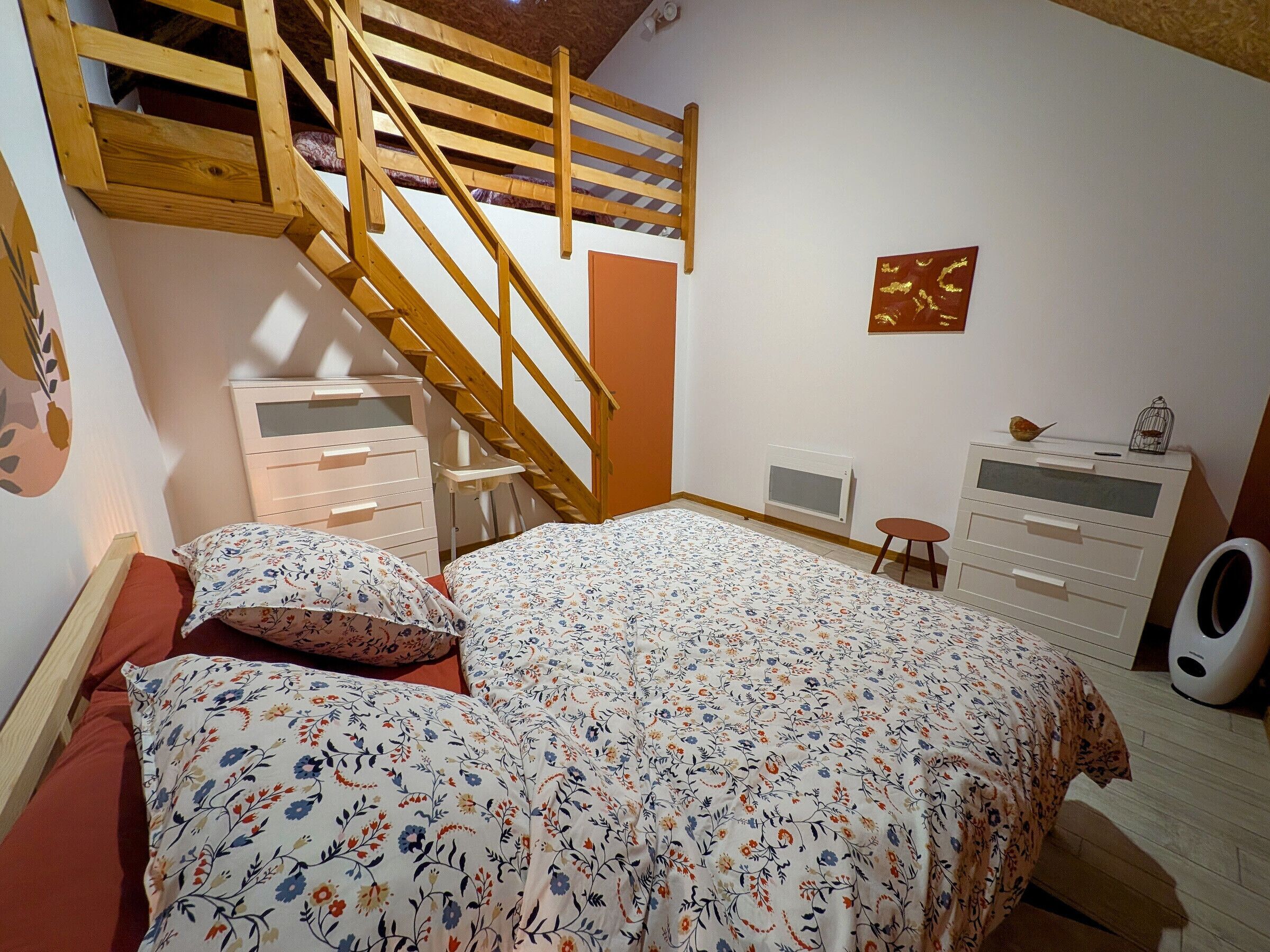 Un dormitorio con escaleras de madera, una cama con ropa de cama floral, un aparador, un radiador y un taburete pequeño.