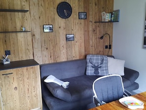 A modern living space with a wooden wall, a black sofa with a deer pillow, a clock, and a small table with a book. - Chalet Waldperle Appartement/Fewo, Dusche, WC, 2-Bettwohnung by Interhome (Arosa)