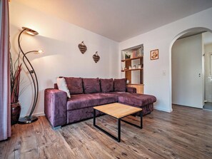 Ett vardagsrum med en lila hörnsoffa, ett soffbord i trä, en golvlampa och en bokhylla.