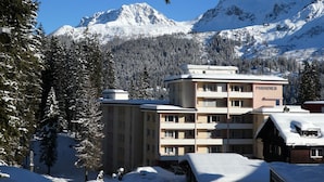 Un immeuble à plusieurs étages, avec une enseigne indiquant « PARADISES » en arrière-plan, entouré d’arbres et de montagnes couverts de neige.