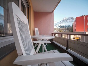 Ein Balkon mit weißem Stuhl und Tisch, Blick auf schneebedeckte Berge und ein rotes Gebäude im Hintergrund.