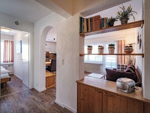 Ein modernes Interieur mit einem Holzschrank, einem Regal mit Büchern und Pflanzen und Blick in eine Küchenzeile.