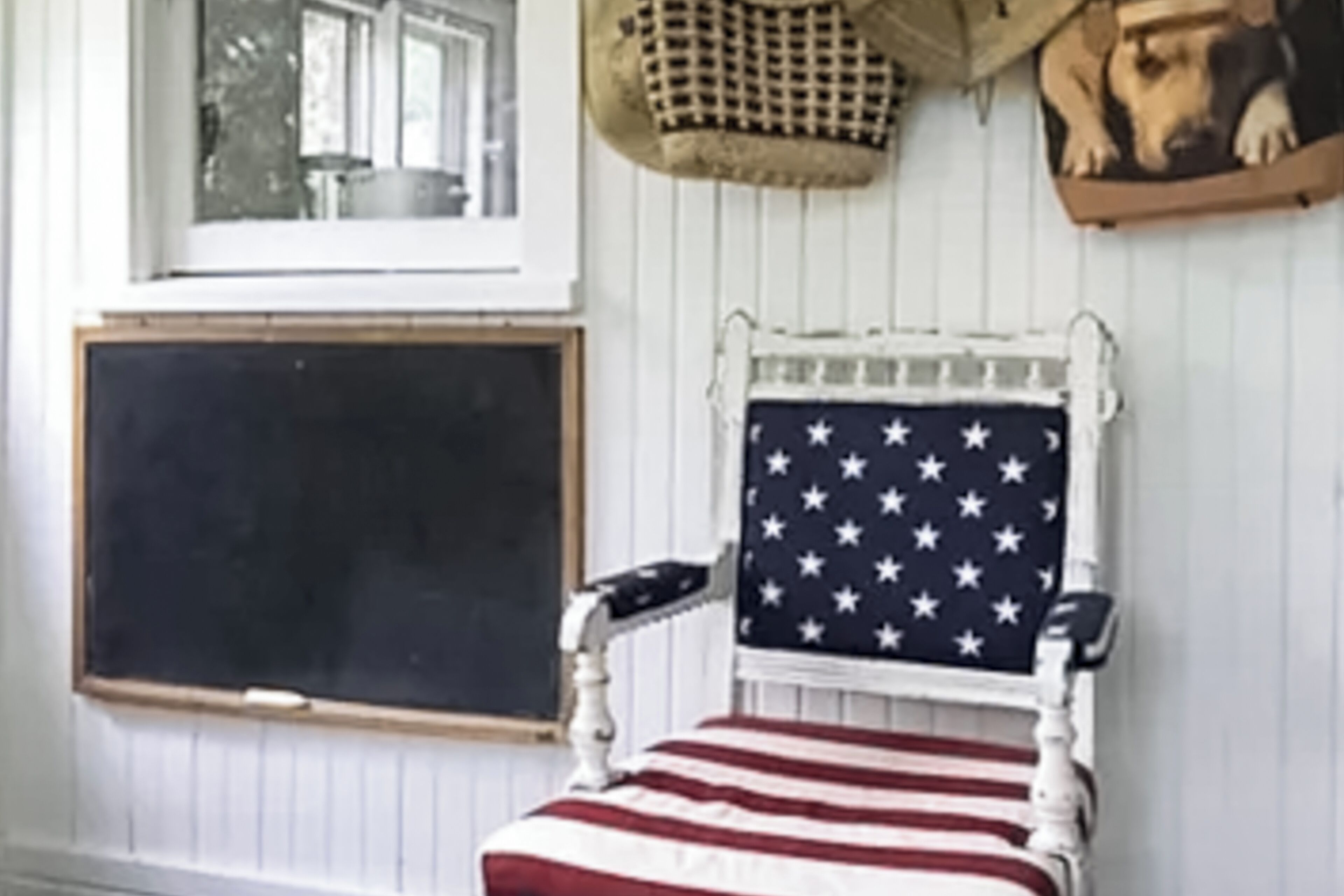 A room with a white wall, a window with a view of trees, a blackboard, and a chair with an American flag cushion.