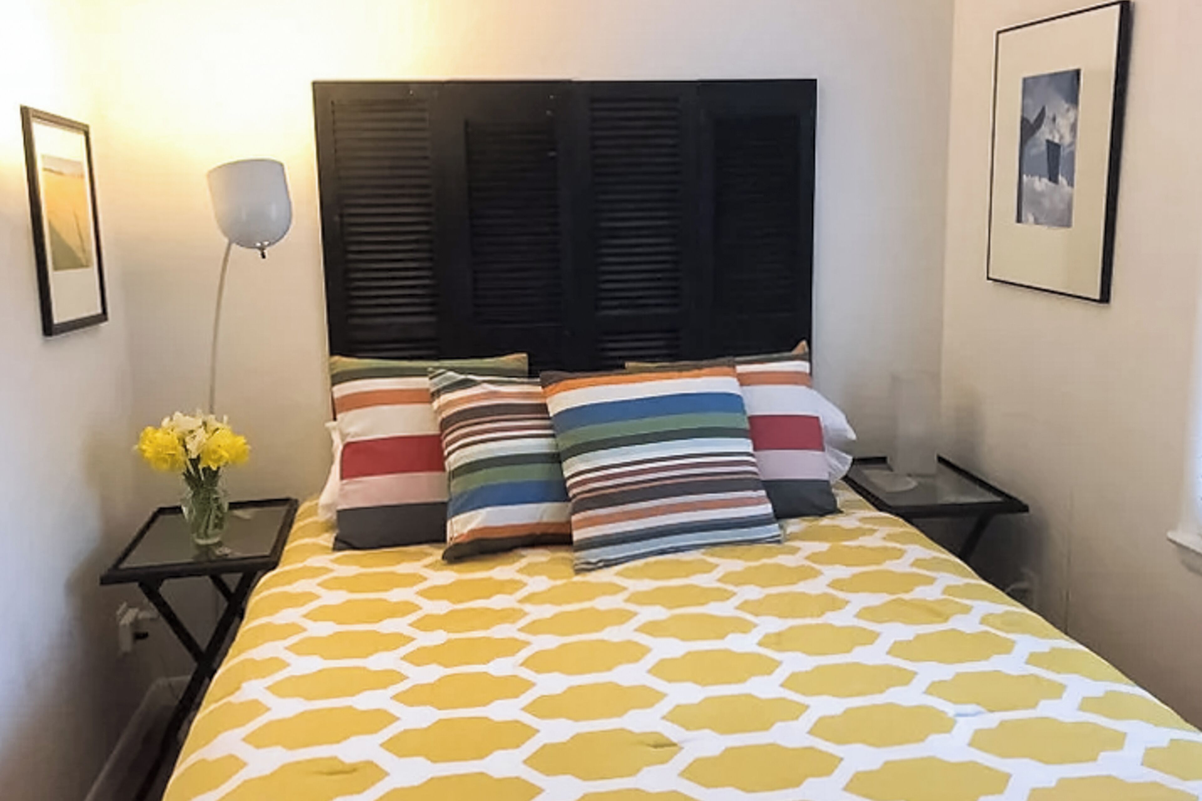 A neatly made bed with a yellow and white patterned bedspread, flanked by two bedside tables with lamps and a vase of flowers.