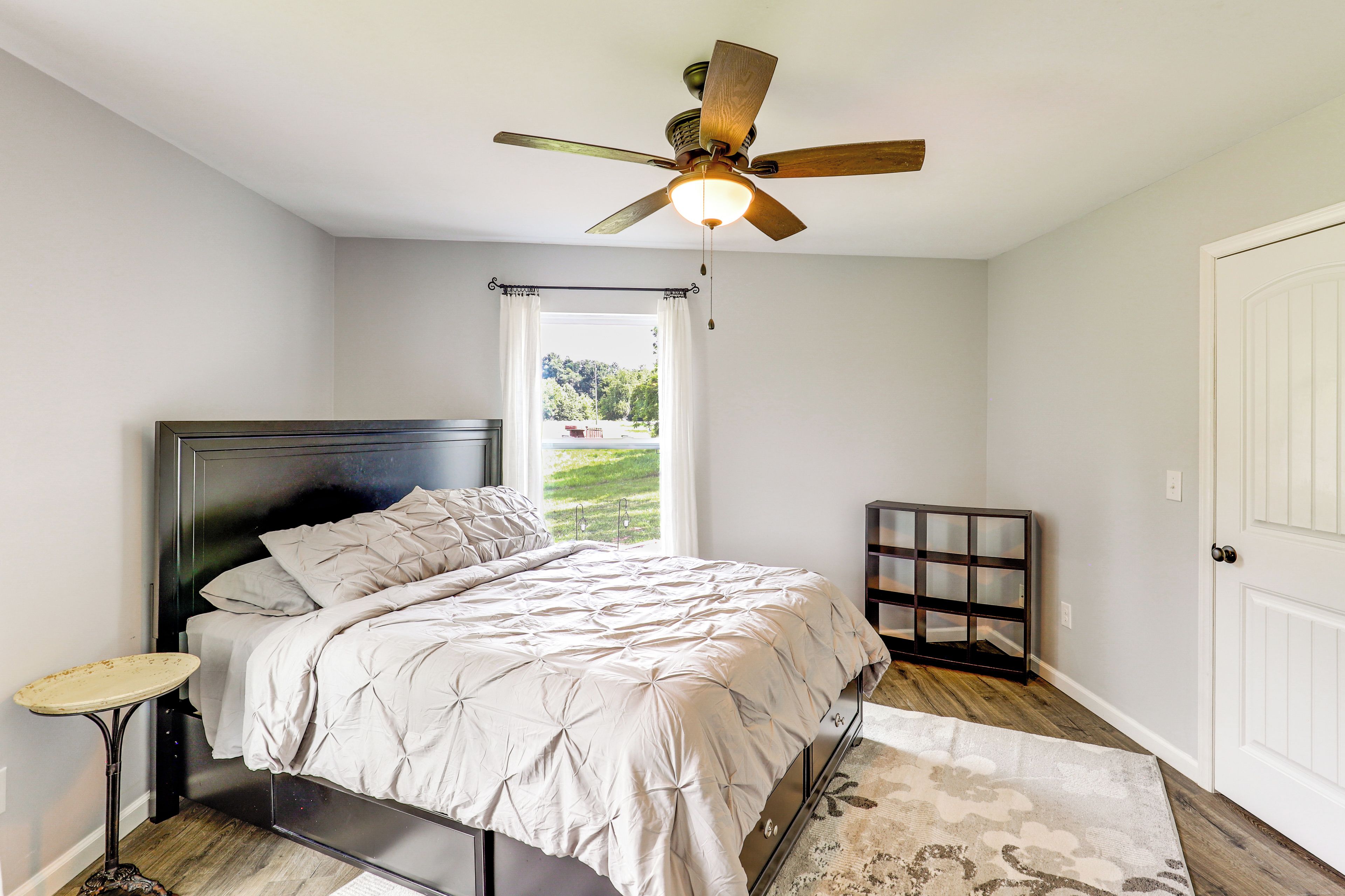 A bedroom with a bed, a ceiling fan, a window with curtains, a door, and a small table.