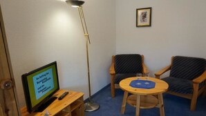 Ein Zimmer mit Fernseher auf einem Holzständer, einer Stehlampe, zwei Stühlen und einem kleinen runden Tisch mit Gläsern.