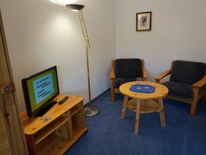 Ein Zimmer mit Fernseher auf einem Holzständer, einer Stehlampe, zwei Stühlen und einem kleinen runden Tisch mit Gläsern.