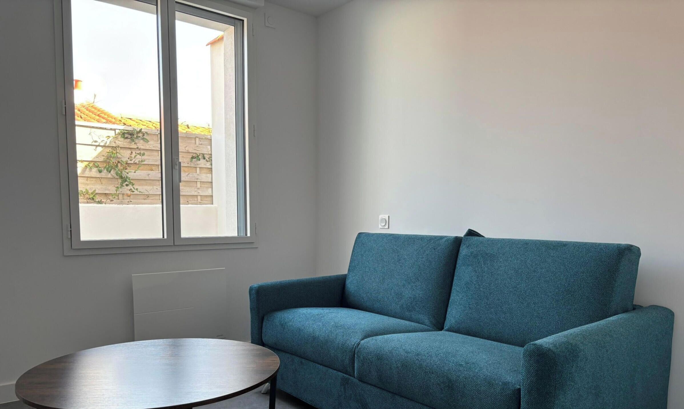 A room with a blue sofa, a small round table, and a window with a view of a building and a fence.