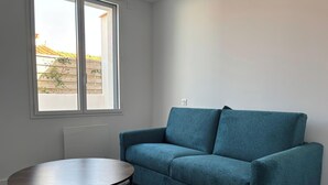 A room with a blue sofa, a small round table, and a window with a view of a building and a fence. - Charming apartment near the beach (Saint-Hilaire-de-Riez)