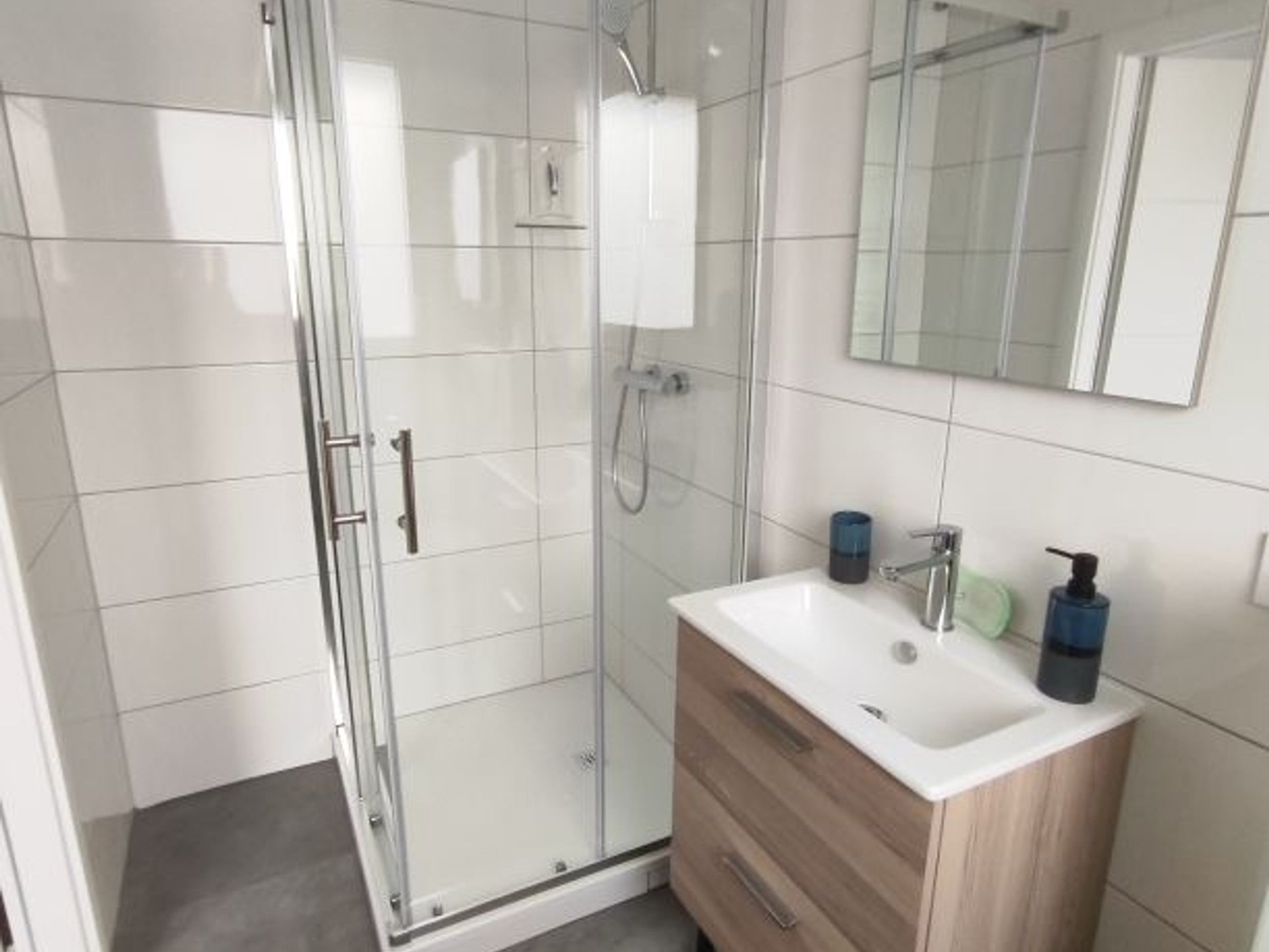 Un bany modern amb una dutxa de vidre, un lavabo blanc i un moble de fusta sota, i un mirall penjat sobre el moble.