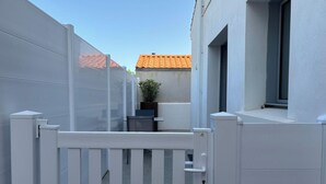 A narrow outdoor corridor with a white gate, a potted plant, and a building with a tiled roof in the background. - Charming apartment near the beach (Saint-Hilaire-de-Riez)
