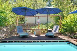 Zona de piscina con dos sillas de descanso y dos sombrillas, rodeada por una terraza de madera y una piscina.