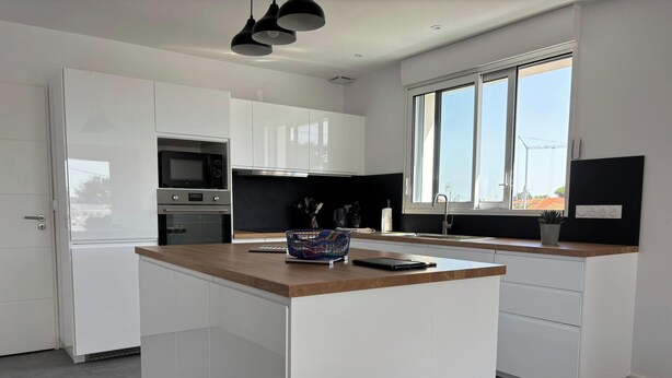 A modern kitchen with white cabinetry, a wooden countertop, and a large window providing natural light. - Apartment near the beach - 6 guests (Saint-Hilaire-de-Riez)