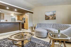 Sala de estar con sofá, reposapiés y una mesita de centro de vidrio, una cocina al fondo y un cuadro en la pared.