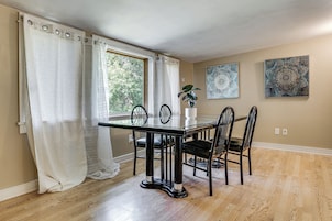 Un comedor con mesa de vidrio, sillas negras, cortinas blancas y dos cuadros colgados en la pared.
