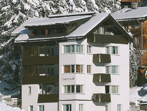 Un hotel de diverses plantes amb balcons, una pista d'esquí al fons i arbres coberts de neu.