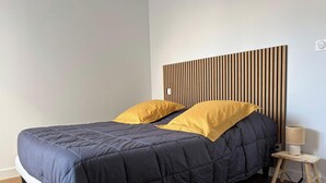 Ein Bett mit dunkelblauer Steppdecke und zwei gelben Kissen, ein Kopfende aus Holzlatten, ein kleiner Nachttisch mit Lampe und ein Lichtschalter an der Wand.