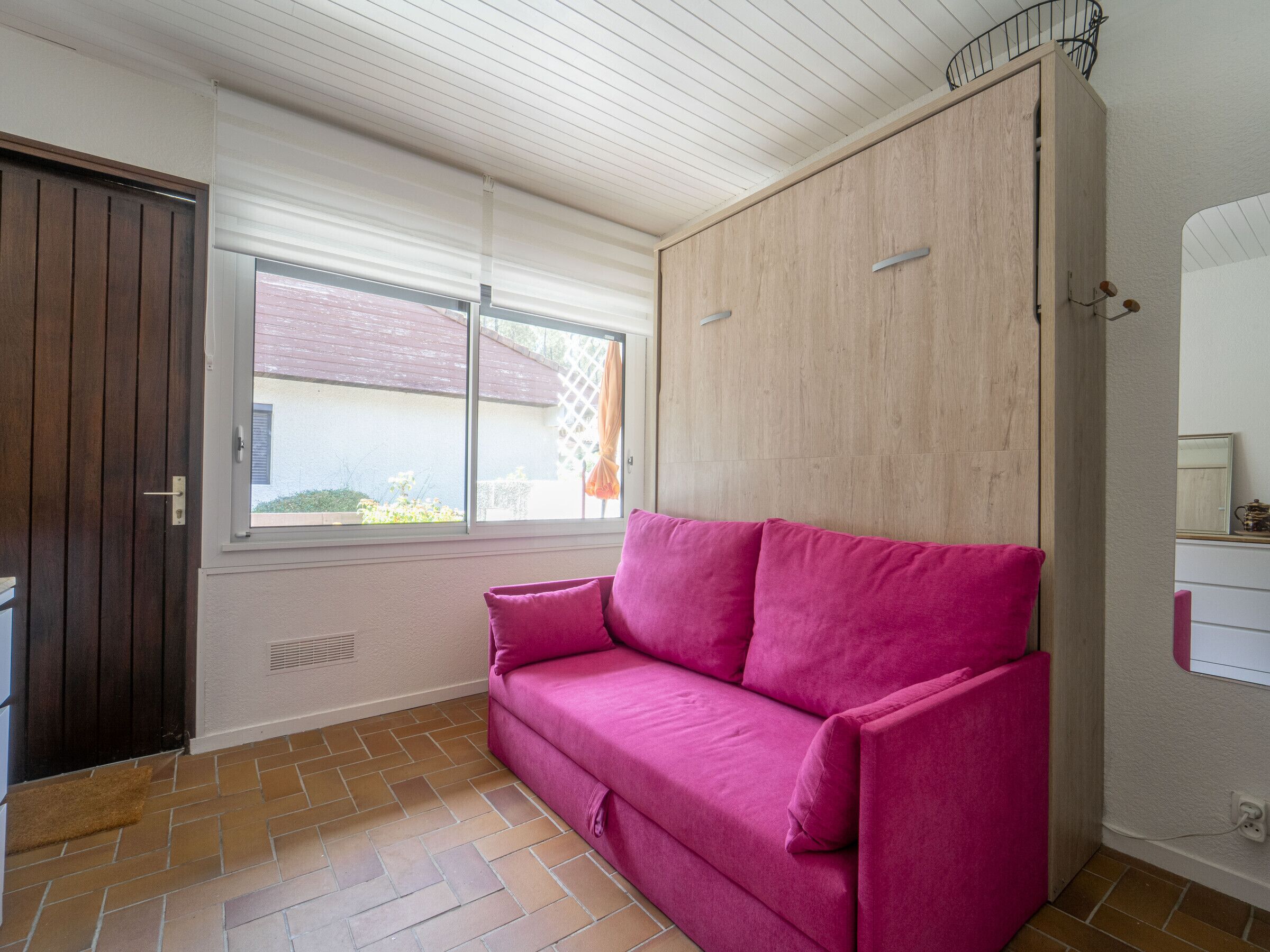 A room with a wooden wardrobe, a pink sofa, a window with blinds, and a tiled floor.