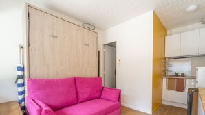 A room with a pink sofa, a wooden wardrobe, a kitchen area with a sink and cabinets, and a tiled floor. - Charming studio at forest edge - 2 guests (Lit-et-Mixe)