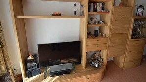 A room with a TV on a glass stand, wooden shelves with books and toys, and a wooden cabinet with glass doors. - Gentiana C-7 Appartement/Fewo, Dusche und Bad, WC, 2-Bettwohnung by Interhome (Arosa)