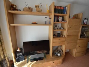 A room with a TV on a glass stand, wooden shelves with books and toys, and a wooden cabinet with glass doors. - Gentiana C-7 Appartement/Fewo, Dusche und Bad, WC, 2-Bettwohnung by Interhome (Arosa)
