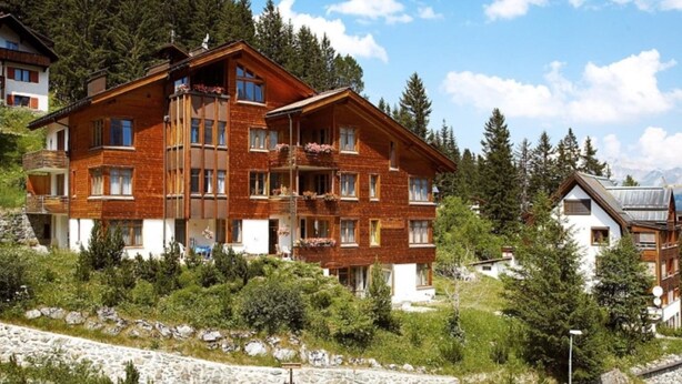 Un hotel de varios pisos con balcones, rodeado de árboles y montañas.