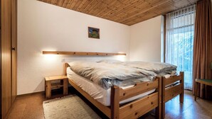 Un dormitorio con una cama de madera, una mesita de noche, un taburete pequeño de madera, un estante montado en la pared y una ventana con cortinas.