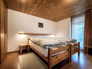 A bedroom with a wooden bed, a nightstand, a small wooden stool, a wall-mounted shelf, and a window with curtains. - Tschuggenweg 2 Appartement/Fewo, Bad, WC, 2-Bettwohnung 2-Bettwohnung by Interhome (Arosa)