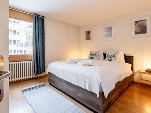 A modern bedroom with a wooden bed, white bedding, and a view of a building through a window with blue curtains. - SKA Haus Appartement/Fewo, Dusche und Bad, WC, 6-Bettwohnung by Interhome (Arosa)