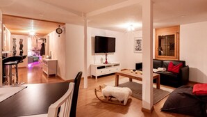 Una sala de estar moderna con un televisor de pantalla plana, un comedor con mesa blanca y una zona de estar acogedora con sofá y reposapiés.