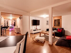 Ein modernes Wohnzimmer mit Flachbildfernseher, einem Essbereich mit weißem Tisch und einer gemütlichen Sitzecke mit Sofa und Hocker.