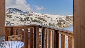 Eine Holzterrasse mit Tisch und Stuhl, mit Blick auf eine gebirgige Landschaft mit schneebedeckten Hängen und blauem Himmel.