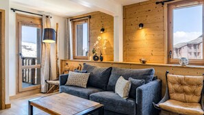Ein gemütliches Wohnzimmer mit einem dunkelgrauen Sofa, einem Holztisch und einem Ledersessel. Zwei Fenster mit Holzrahmen, eine Holzbalkendecke und eine kleine Regal mit Dekorationen.