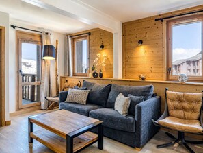 A cozy living room with a dark grey sofa, a wooden coffee table, and a leather armchair. There are two windows with wooden frames, a wooden ceiling, and a small shelf with decorative items. - Spacious apartment, close to slopes - 6 guests (Belle Plagne)