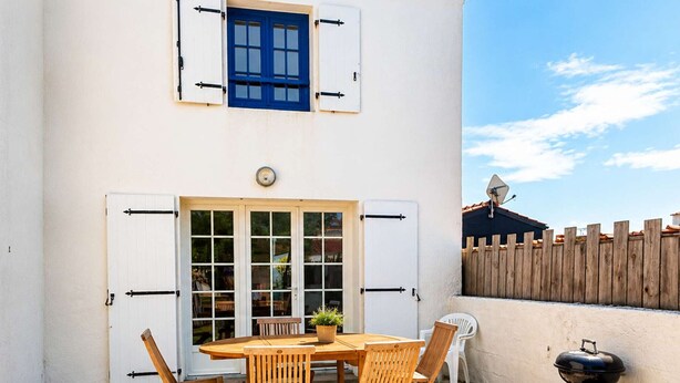 Eine Terrasse mit Holztisch und -stühlen, einem Grill und einem weißen Gebäude mit blauem Fenster und Fensterläden.