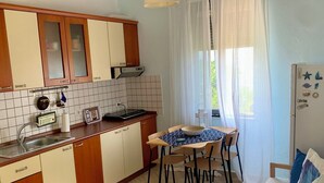 Ett kompakt kök med en matplats, ett kylskåp med magneter och ett fönster med tunna, genomskinliga gardiner.