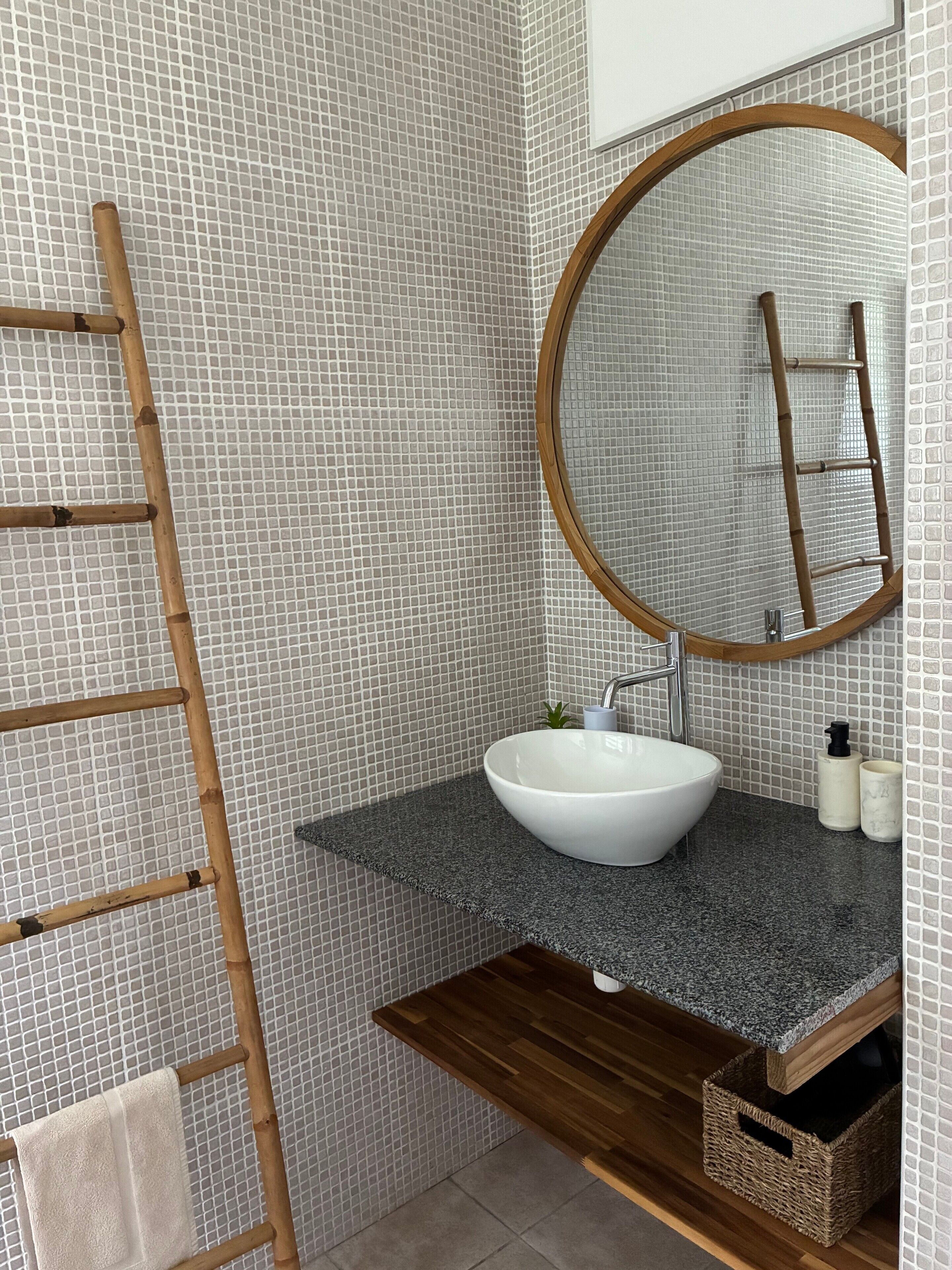 A bathroom with a white sink, a round mirror, a wooden ladder, and a towel hanging on the wall.