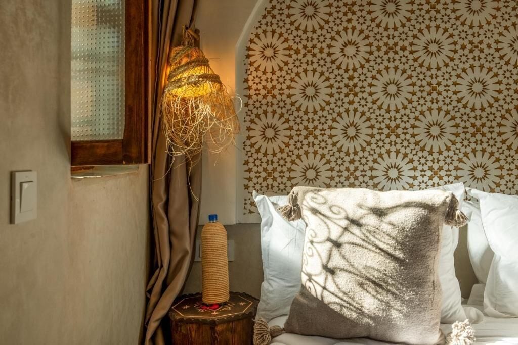 A room with a patterned wall, a bed with white pillows, a wooden nightstand with a lamp, and a window with curtains.