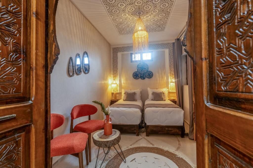 Una habitación con dos camas, una silla roja, una mesita con un florero y un techo decorativo.