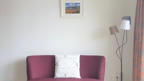 Ein Zimmer mit einem roten Sofa, einem weißen Kissen mit Naturmuster, einem gerahmten Landschaftsbild an der Wand und einer Stehlampe.