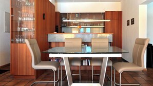 Uma sala de jantar moderna com mesa de vidro, cadeiras de metal, armário de madeira com prateleiras de vidro e uma cozinha ao fundo.